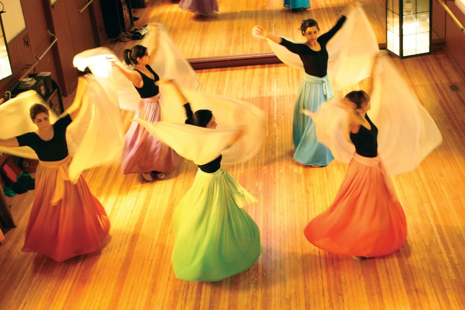 Dancers in flowing skirts and white veils turning on a wooden studio floor.