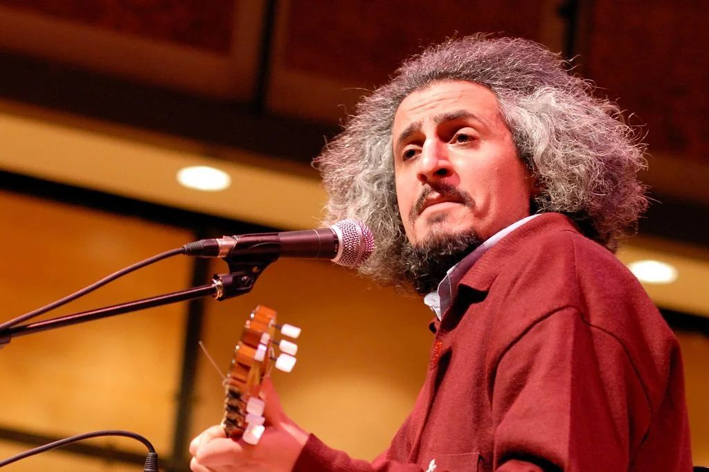 Composer Mohsen Namjoo singing into a microphone, a guitar neck in the foreground.