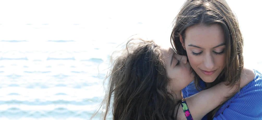 Still from The Neighbor: a woman holds a young girl against a bright seascape.